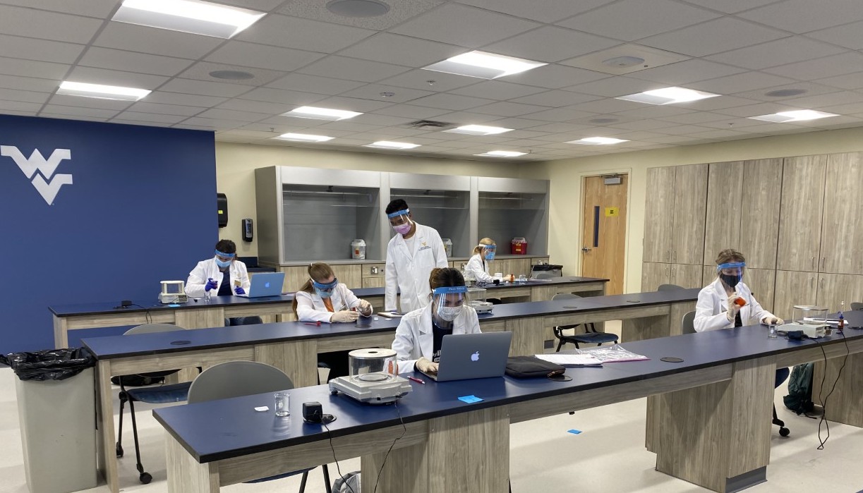 Students in white coats and personal protective gear work in a lab.