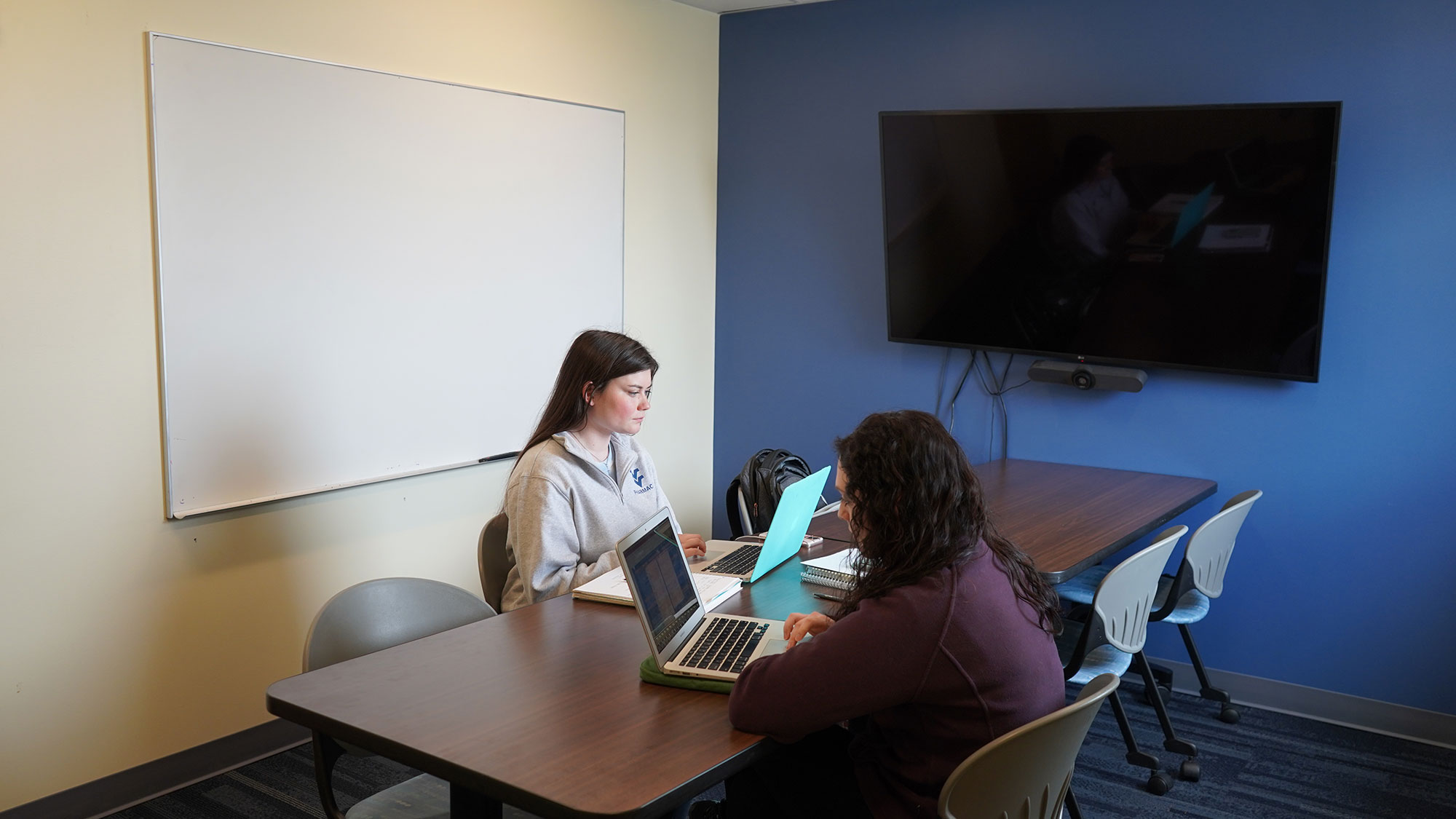 Two students show seated at a table in the middle of a smaller room with a monitor and white board present.