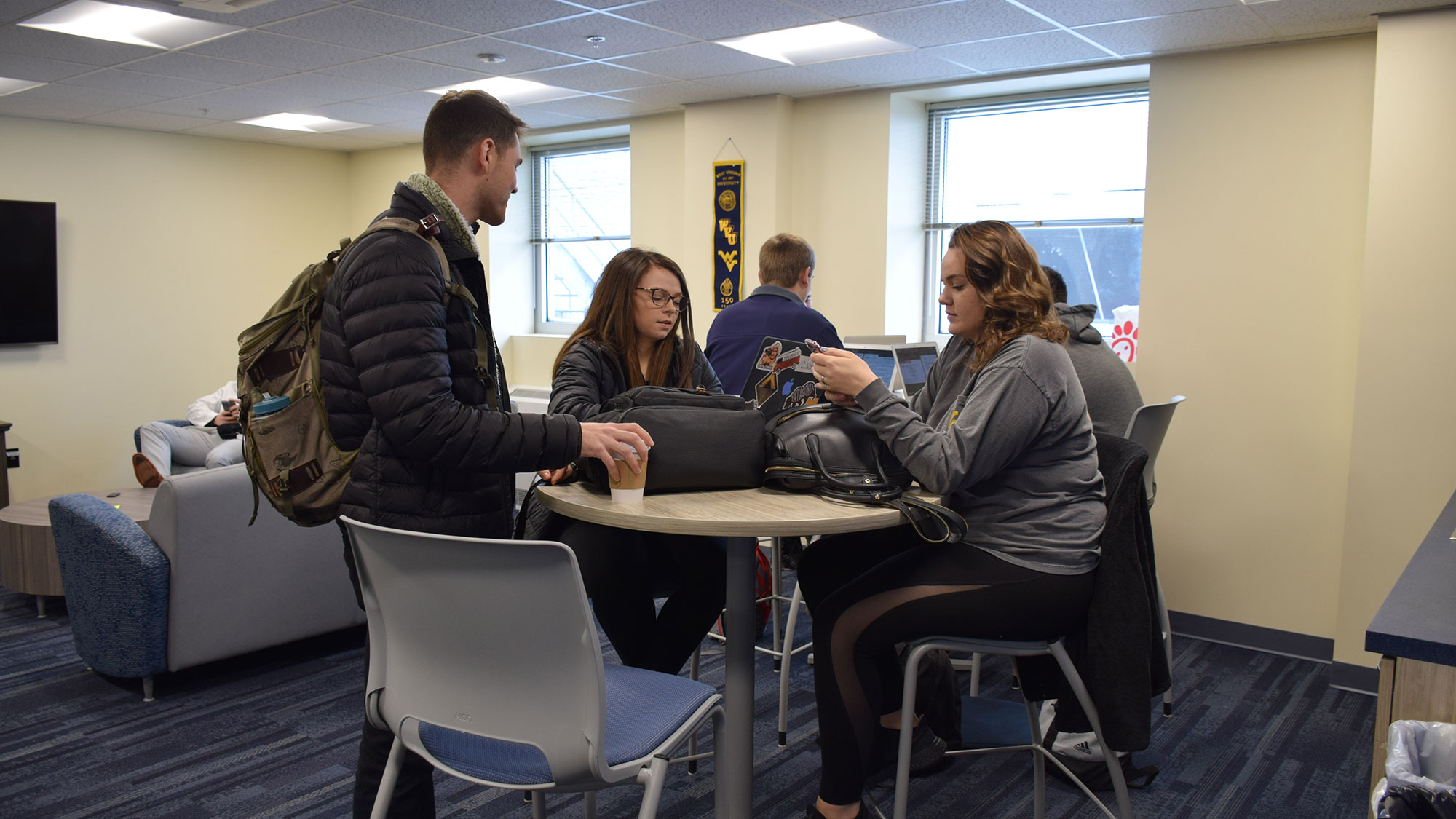 A number of students are seen relaxing in the lounge room.  There are tables in the center and a couch at the rear along with other amenities.