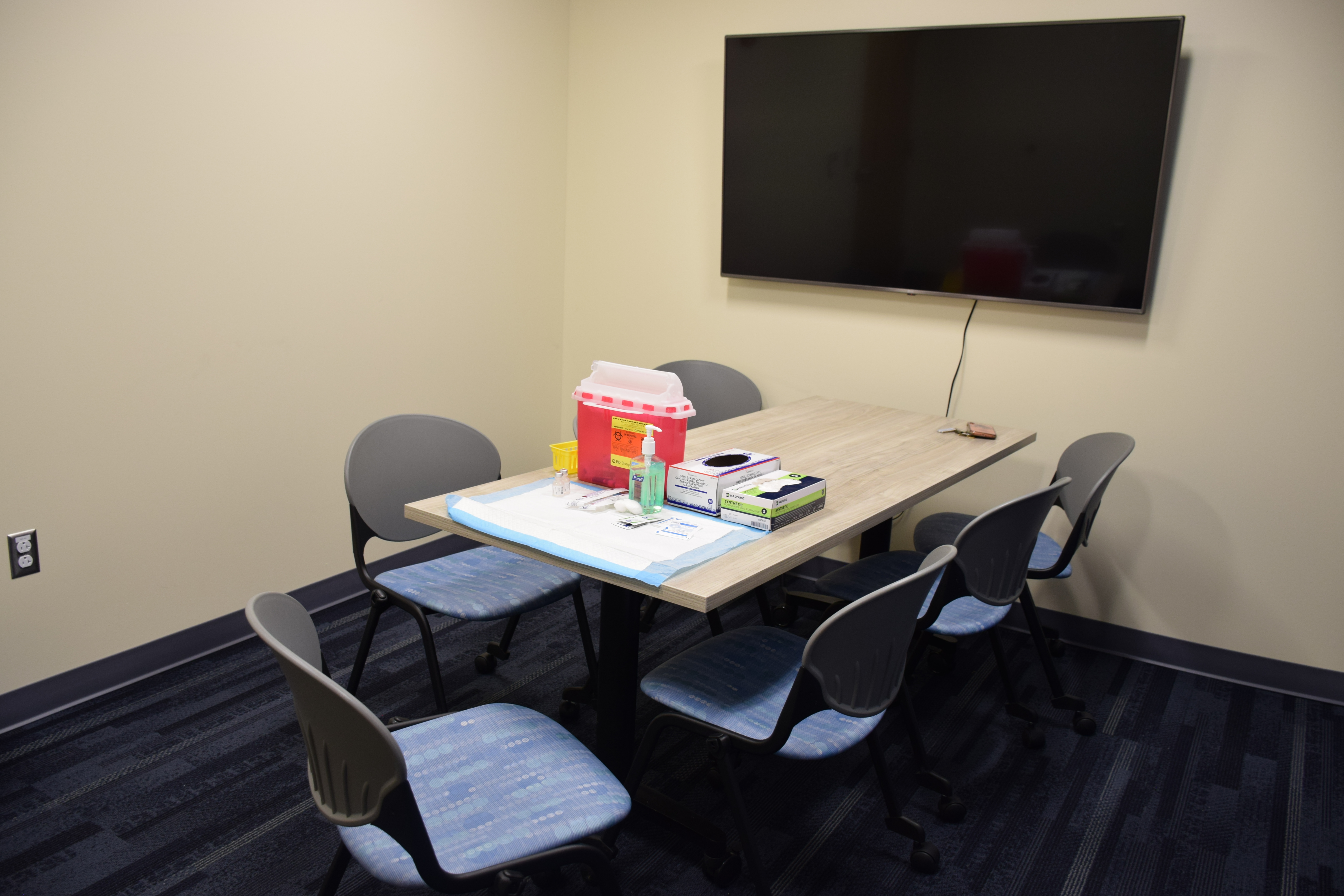 A table in a yellow room with a tv screen on the wall and immunization supplies on the table.