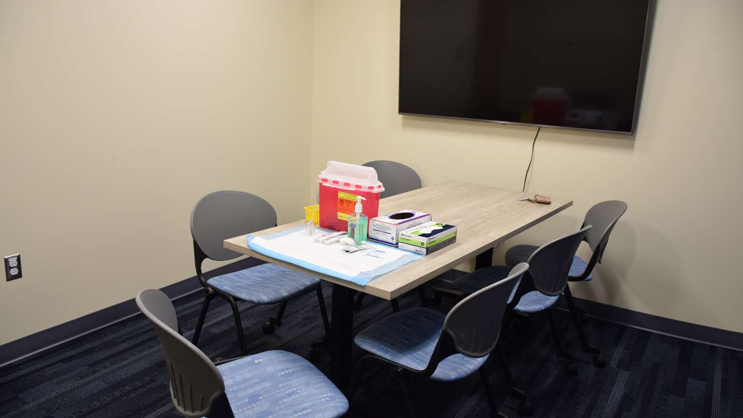 A table in a yellow room with a tv screen on the wall and immunization supplies on the table.