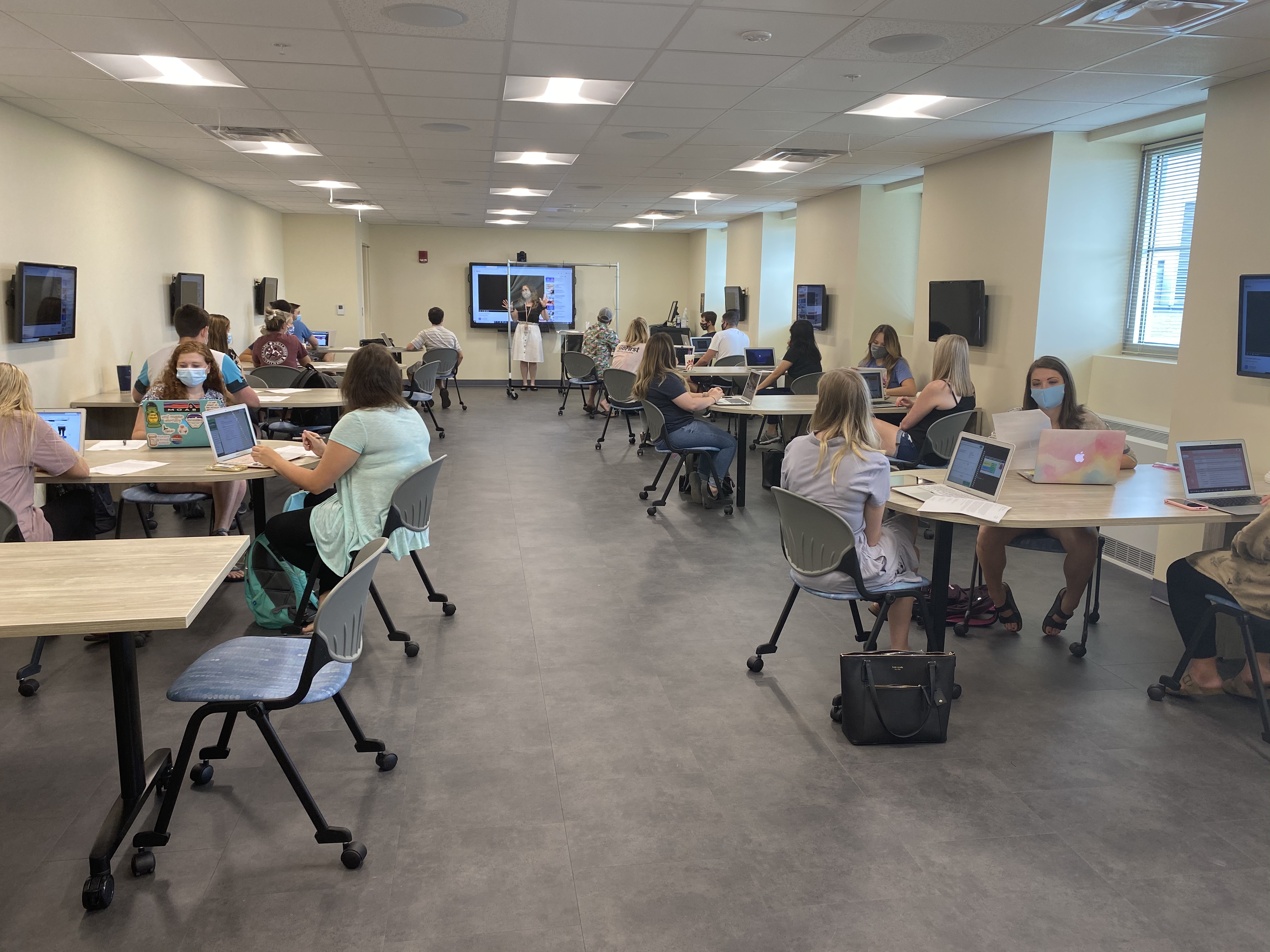 Students sit a tables in a long classroom with computers ad windows.