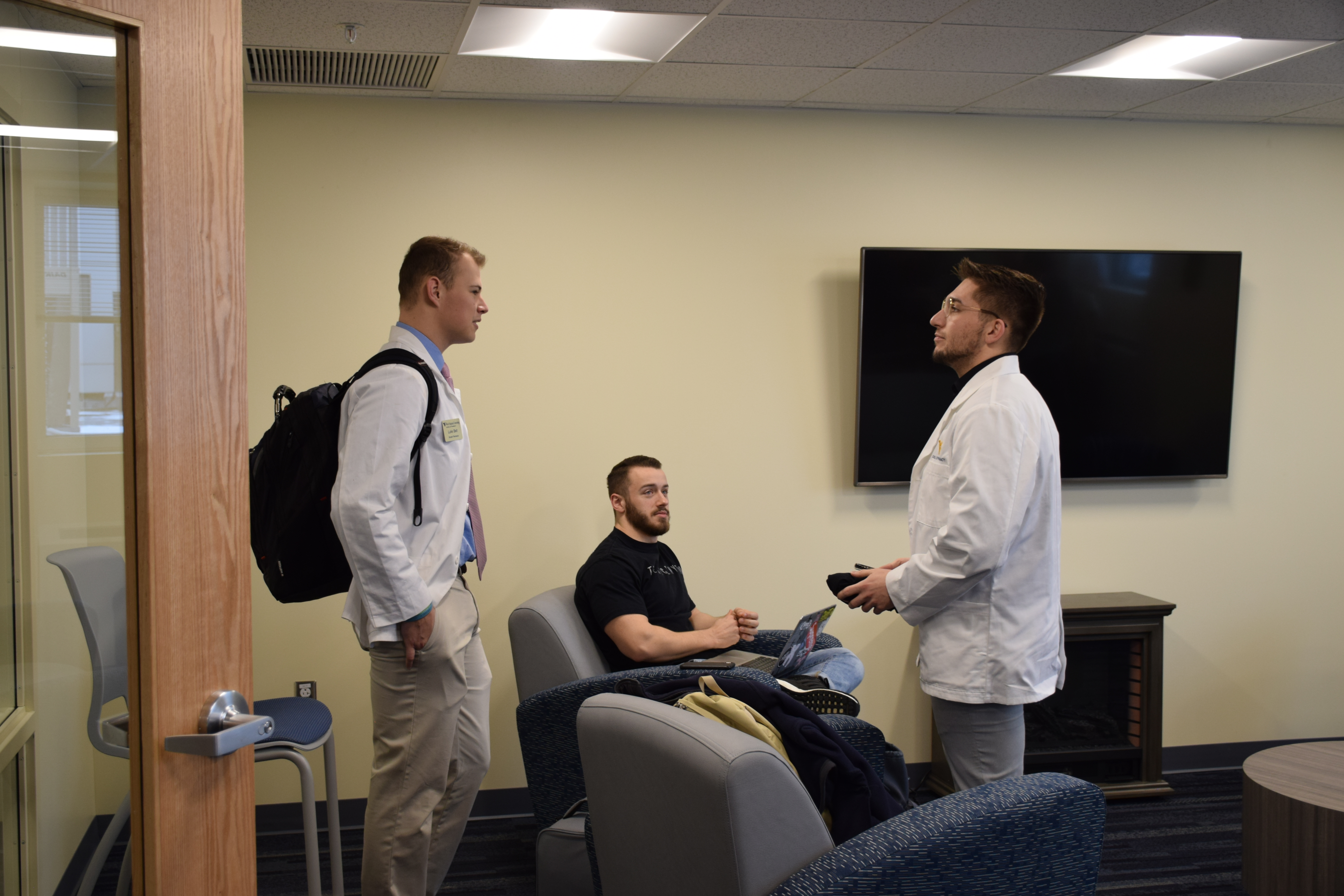 Three male students wearing white coats talk in the student lounge.