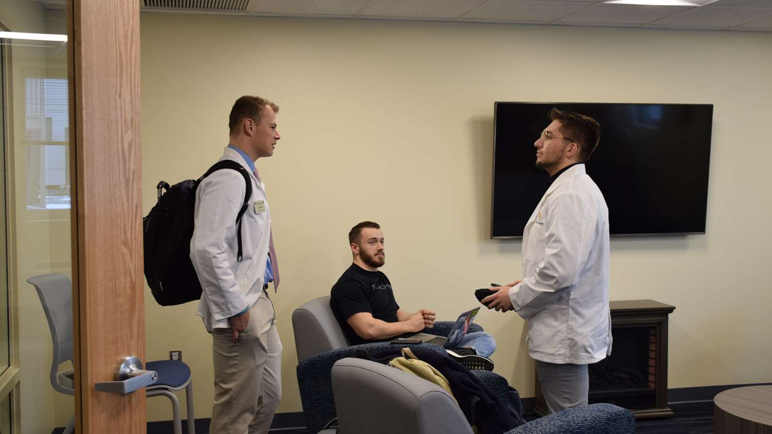 Three male students wearing white coats talk in the student lounge.
