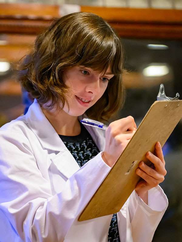 A pharmacy Student in a white coatd holding a clipboard and taking notes.
